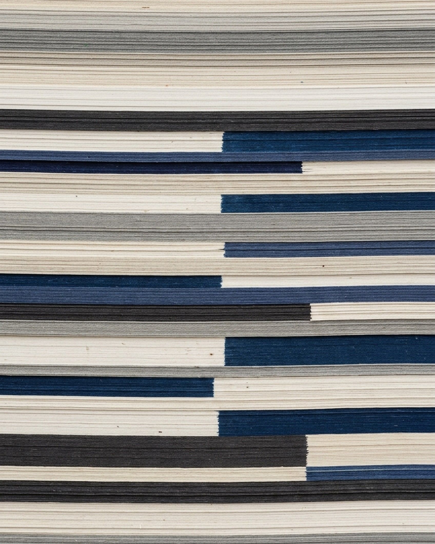 A tall stack of paper and card seen on its cut edge: fine horizontal strata in off-white, grey, near-black and deep blue, offset in stepped blocks.