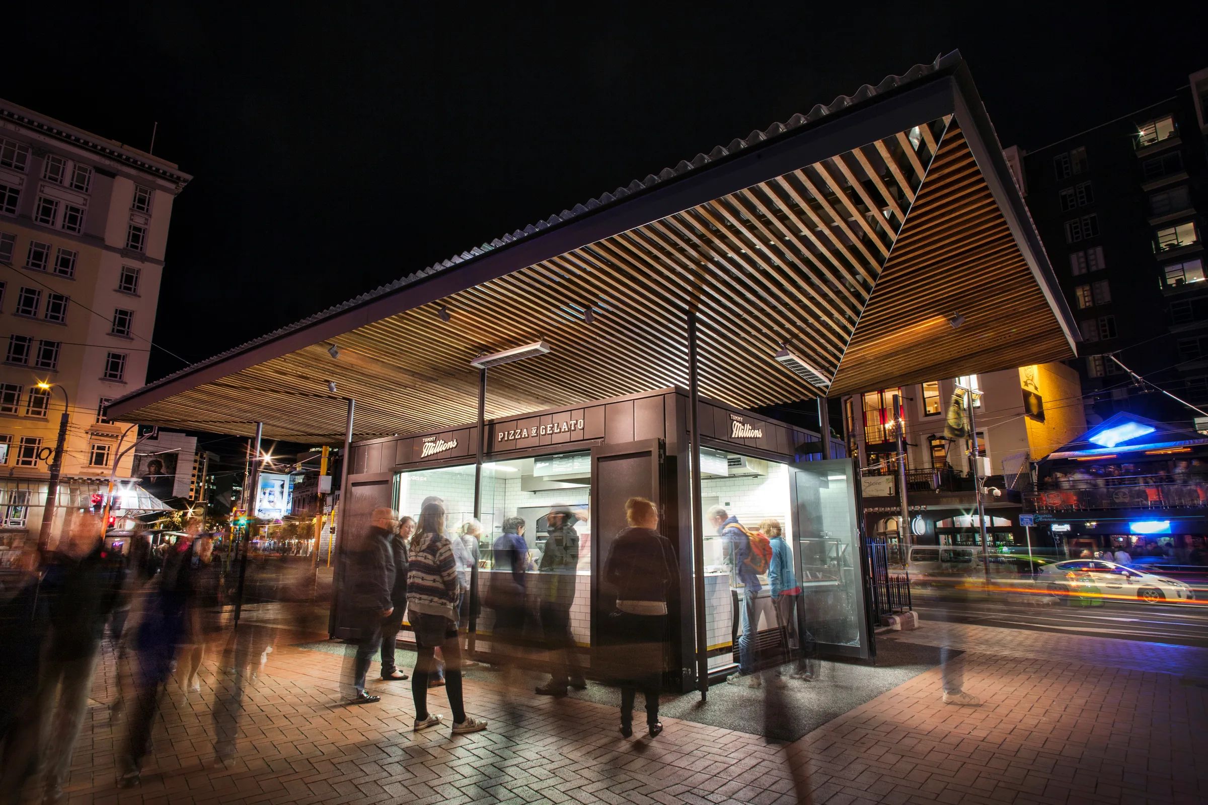 A timber-slatted kiosk lit from within at night on a city corner, the figures of passers-by blurred by a long exposure.